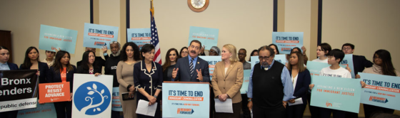 Large number of people near podium, most holding signs about ending immigrant criminalization.