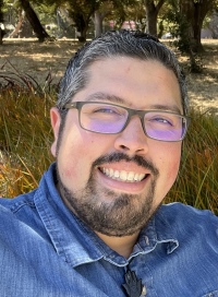Jaime Perez Headshot Image features Jaime in the forefront wearing a blue jean shirt, a forrest in the backdrop.
